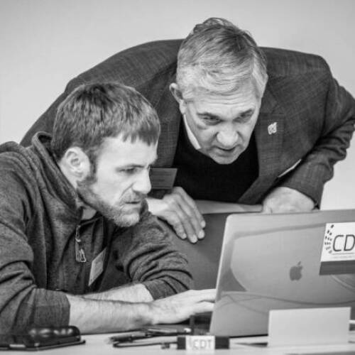 A black and white image of two men one standing and the other sitting both look closely at a laptop